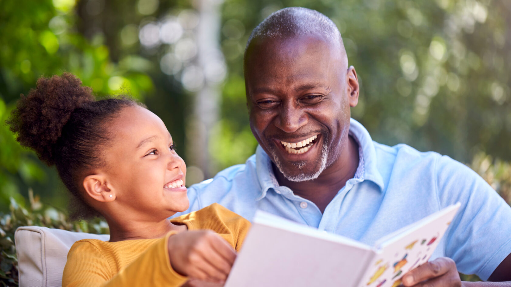 Una nieta le lee un libro a su abuelo al aire libre.