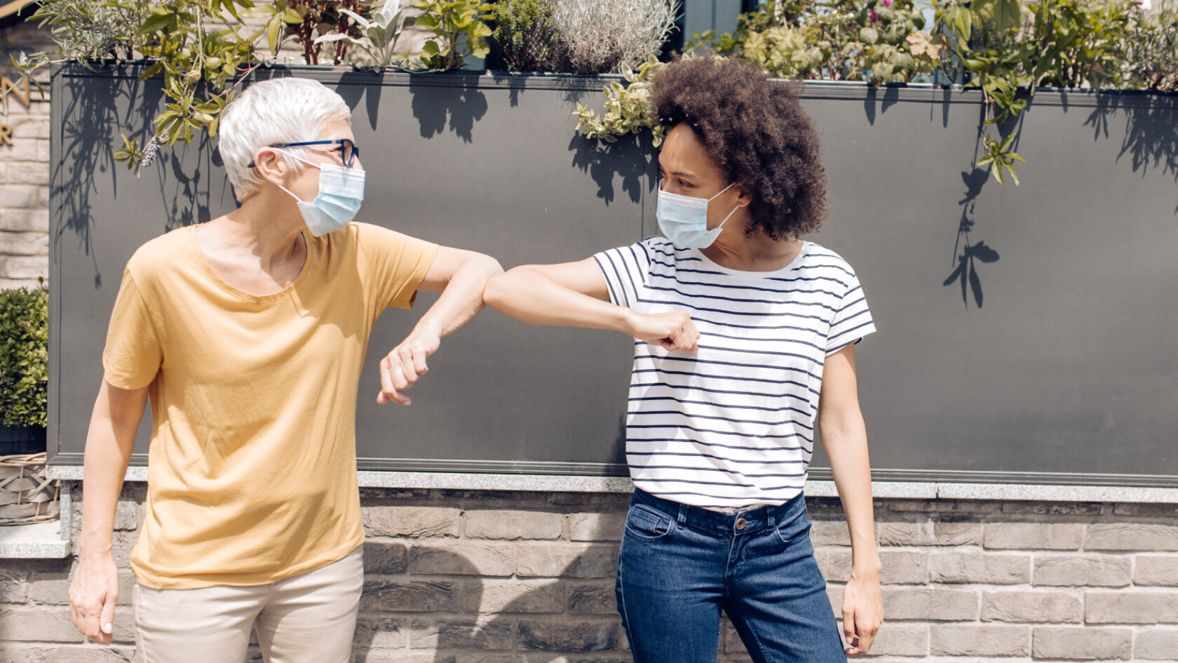 Friends wearing face coverings tap elbows to greet each other outside.